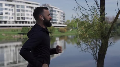 Adult Man Runs in the Park Near the City Camera Following Runner in Wireless Headphones Healthy