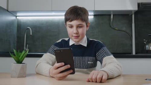 Boy Using Smartphone Sitting at Kitchen Table Indoors