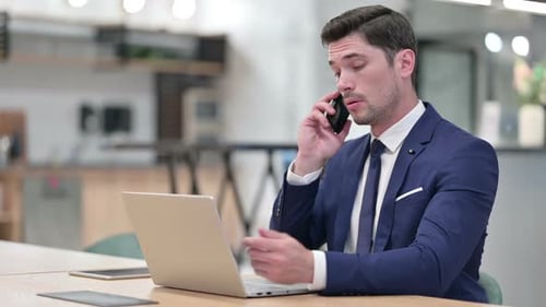 Businessman with Laptop Talking on Smartphone in Office