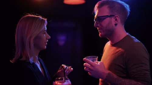 Adult Couple Drink Cocktails in the Bar. Man in Glasses and Blond Woman Talking at the Disco