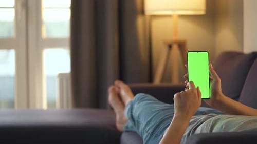 Woman at Home Lying on a Sofa and Using Smartphone with Green Mock-up Screen in Vertical Mode. Girl