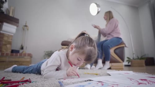 Close-up of Cute Brunette Girl Drawing at the Foreground. Blurred Young Man and Woman Talking and