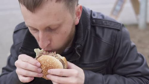 Young Man with Beard in the Street Cafe Biting Tasty Big Burger with Cheese