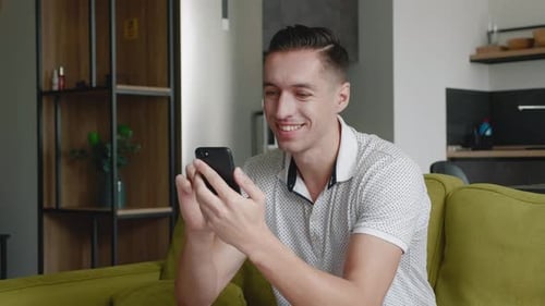 Close Up of Cheerful Young Man Sitting on the Sofa Using Smartphone Smile in the Modern Apartment