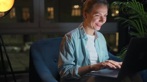 Smiling Woman Typing on Laptop in Armchair at Night
