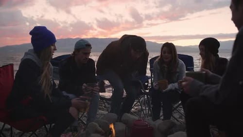 Group of people holding mugs around campfire as man checks kettle