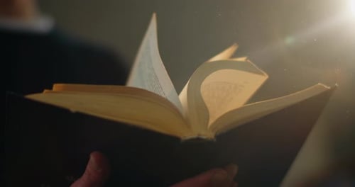 Close-up, the Guy Is Quickly Flipping an Old Book and Dust From It Is Flying Out To the Light. A