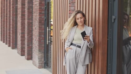 Woman in Suit Leaning Against Building with Smartphone