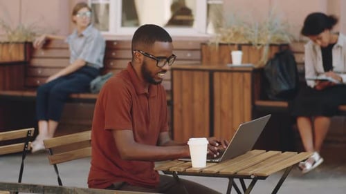 Afro-American Man Working on Laptop at Table in Outdoor Street Cafe