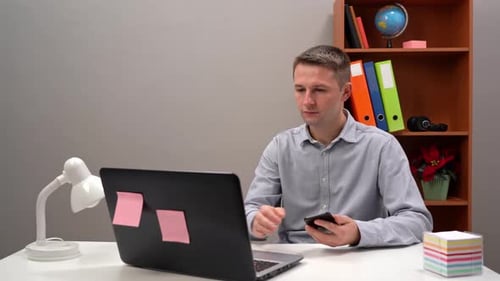 Young Guy Office Worker Manager is Typing Text in the Phone and in the Computer