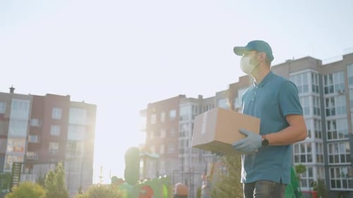 The Male Deliveryman in Masks and Protective Gloves Carries a Parcel to the Customer with a Terminal