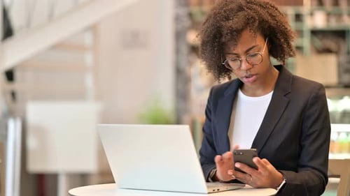 Hardworking African Businesswoman Using Smartphone and Laptop at Cafe