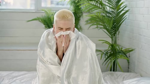 Young Adult with Cold Sits with Blanket on Bed
