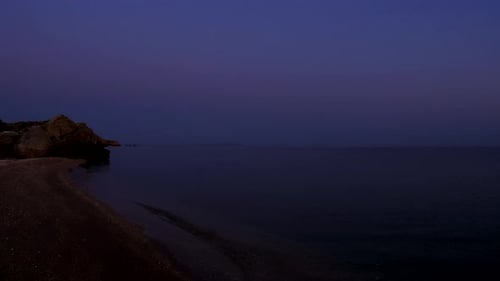 Morgenröte. Meereswellen waschen den Sandstrand. Eine felsige Klippe ragt ins Meer hinein. Der Himmel wechselt seine Farbe