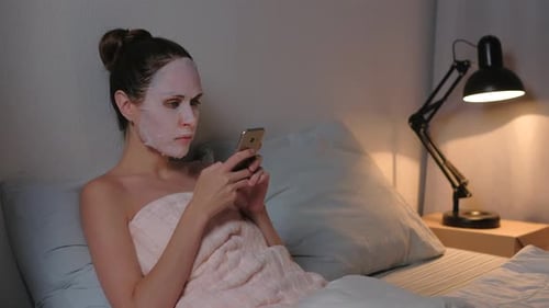 Woman with Face Mask Using Cell Phone in Bed