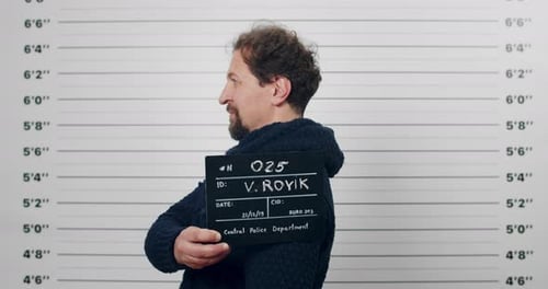 Portrait of Male Adult Criminal with Beard Turning to Sides While Holding Sign for Photo in Police