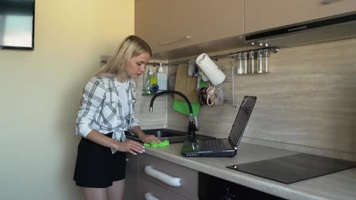 Woman Works on Laptop While Cleaning Kitchen