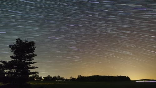 startrails over rural landscape with field in front of forest sihlouette