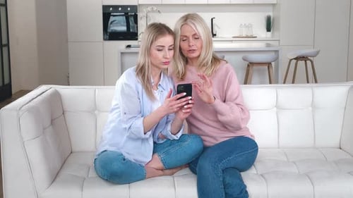 Two blonde women look at a phone on couch