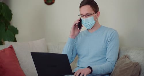 Man Working on Laptop, Talking on Smartphone, Wearing Mask