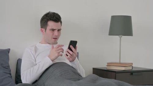 Man Reacts in Disbelief to Smartphone in Bed