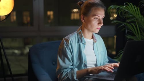 Woman is Sitting in the Armchair and Working on a Laptop at Night