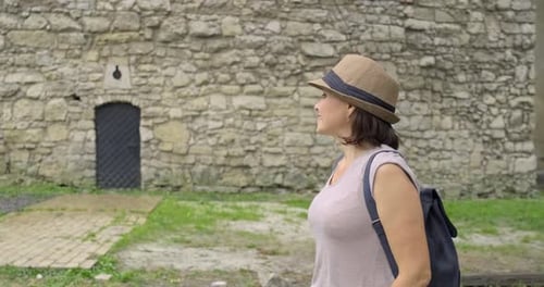 Woman Tourist in Hat with Backpack Walking in the Park Near Ancient Medieval Castle