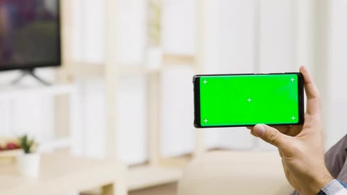 In Modern and Bright Living Room Man Holds Horizontally a Green Screen Phone in His Hand