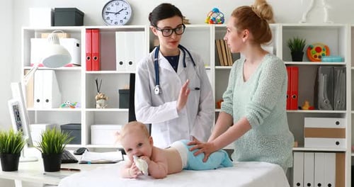 Mutter und Arzt untersuchen das auf dem Tisch liegende und spielende Baby und besprechen die Behandlung.