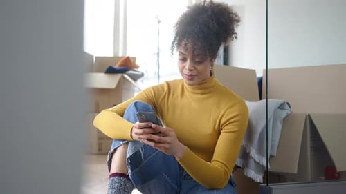 Young Woman Using Mobile Phone In Lounge Of New Home On Moving Day Surrounded By Removal Boxes