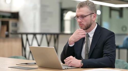 Pensive Young Businessman Thinking and Working on Laptop in Office
