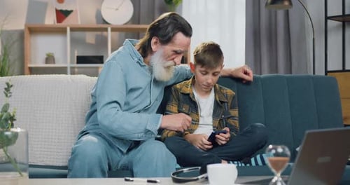 Niño sentado en el sofá en una habitación acogedora junto con su abuelo barbudo y usa un teléfono inteligente