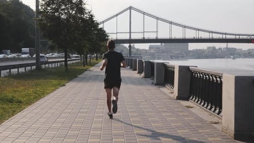 Young fit athlete running along city river at promenade towards bridge at sunset wearing black