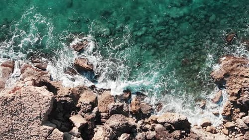 Aerial top view of sea coast line with ocean waves hitting rocky seashore creating white foam