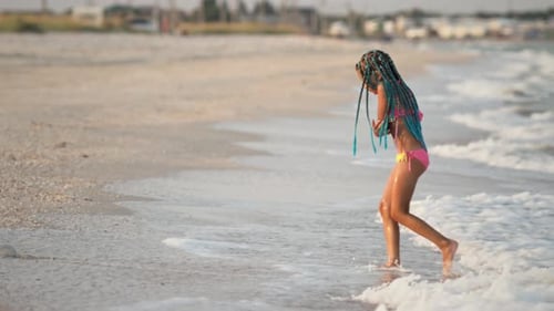 A Girl with African Braids in a Summer Costume Plays on the Beach with Shells Near the Sea with
