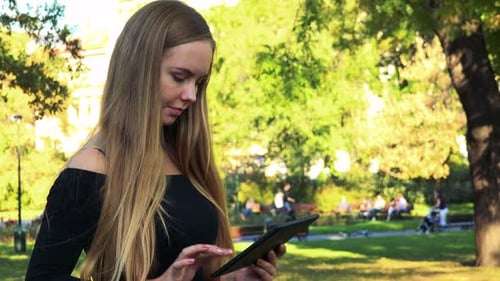 Young Beautiful Woman Works on Tablet in the Park in Sunny Day