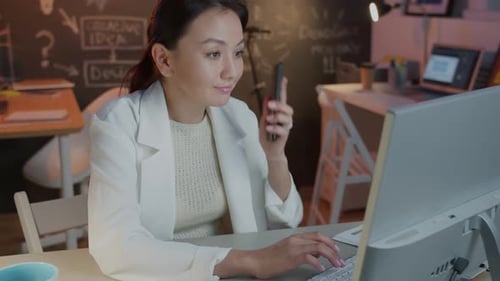 Cheerful Young Businesswoman Working with Computer and Speaking on Mobile Phone in Dark Office Late