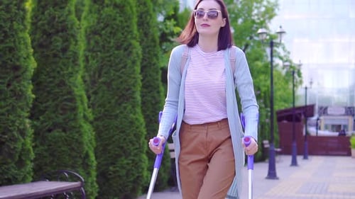 lBeautiful Stylish Positive Young Woman with Injury on Crutches Walking Down the Street Sunny