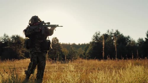 Camouflaged Figure Walks With Rifle in Sunset Field