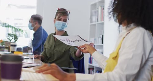 Woman wearing face mask passing a document to her colleague at office