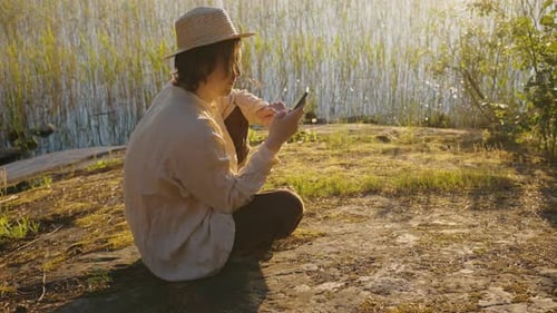 Man in Hat Tries to Get Cell Signal on Phone on River Bank
