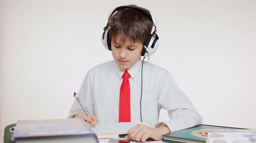 Young Concentrated Caucasian School Kid in Red Tie Sitting at Table Wiritng Listening Music Through