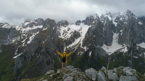 A Hiker Stands on Top of a Mountain and Looks at Snow-capped Mountains, Raising His Hands in Victory