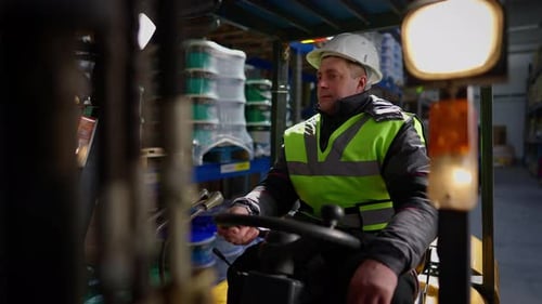 Male Caucasian Forklift Driver Looking Around at Freight on Racks in Warehouse Sitting in Vehicle