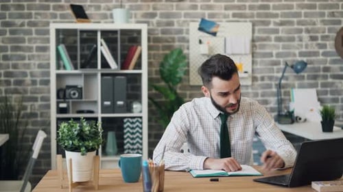 Young Man Writing in Notebook and Using Laptop Sitting at Desk in Office