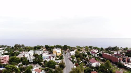 Aerial View of Rooftops Among Trees on the Coast and a Lonely White Yacht in the Blue Sea on a Sunny