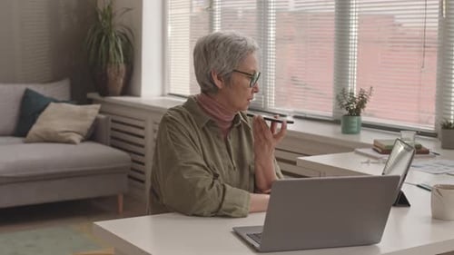 Mature Woman Uses Smartphone at Laptop in Home Office
