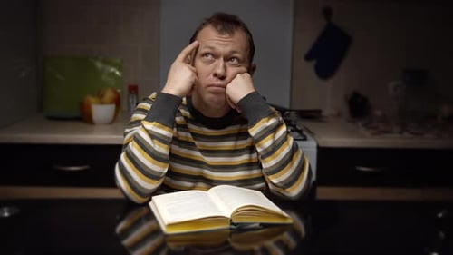 Student guy sitting thinking over a textbook while sitting in the evening at the kitchen