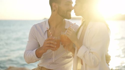 Smiling Hispanic Couple with Cocktails Kissing Against Sea