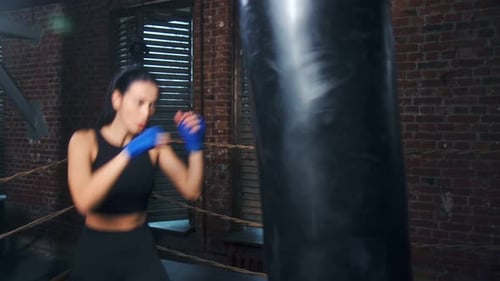 Fierce Asian Fit Woman with Wrapped Hands Practicing Kickboxing on Punching Bag at Box Studio